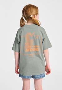 Grön bomullströja med en stor orange grafisk tryck på baksidan med texten "the stadium ANTHEMS" och lekfull design av karaktärer.