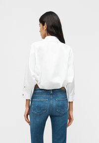 Γυναίκα με μακριά σκουρόχρωμα μαλλιά, που φοράει άνετο λευκό πουκάμισο με κουμπιά και μπλε τζιν, γυρισμένη προς τα πίσω μπροστά σε ένα απλό φωτεινό φόντο.