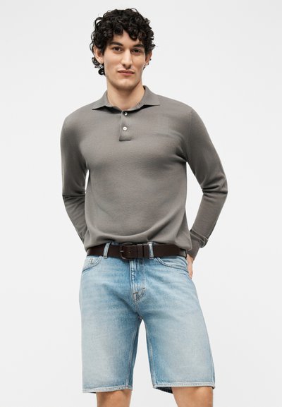 Άνδρας με σγουρά μαλλιά, φοράει γκρι polo με μακριά μανίκια που είναι μισοφεμένο μέσα σε ανοιχτόχρωμα τζιν σορτς με σκούρα ζώνη, στέκεται με τα χέρια πίσω από την πλάτη.