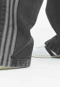 Weiße Sneakers mit Perforationen und schwarzen Akzenten, kombiniert mit dunkelgrauen Jogginghosen, die drei hellgraue vertikale Streifen aufweisen.