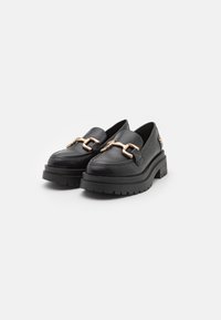 Zwarte leren instappers met een chunky platform, voorzien van een gouden kettingaccent aan de voorkant en een gestructureerde rubberen zool.