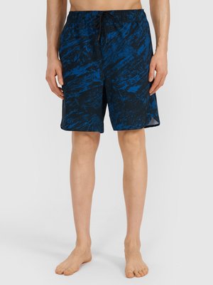 Man draagt knielange zwarte en blauwe zwembroek met patroon en een koord in de taille, staat blootsvoets op een effen achtergrond.