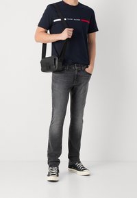 Black crossbody bag with a smooth texture, featuring a front clasp. Worn with dark gray jeans and a navy T-shirt with red and white stripes.