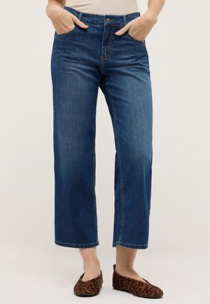 Frau trägt blaue, gerade geschnittene cropped Jeans, beigefarbenes, eingestecktes Oberteil, Hände in den Taschen und Leopardenmuster-Ballerinas, steht vor hellem Hintergrund.