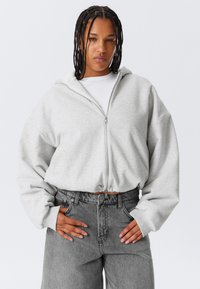 Siva zip-up dukserica s krojenim dizajnom, širokim rukavima i podesivim donjim rubom s vezicama, uparena s visokim sivim trapericama.