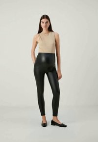 Leggings pretos em couro sintético com uma cintura alta; combinados com um top bege canelado sem mangas. O modelo usa sapatos baixos pretos; textura suave.