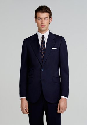 Marineblauwe op maat gemaakte colbert met twee knopen, notched revers, twee voorzakken en een witte pochet. Wordt gekenmerkt door een gestreepte stropdas.