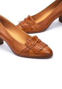 Escarpins en cuir marron avec un talon carré, dotés d'un détail à franges et d'une accent tressée au niveau des orteils. Texture lisse, design élégamment façonné.