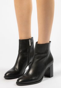 Bottes noires en cuir à chevilles avec un talon bloc haut et un bout pointu. Elles disposent d'une fermeture à zip sur le côté et d'une texture lisse.