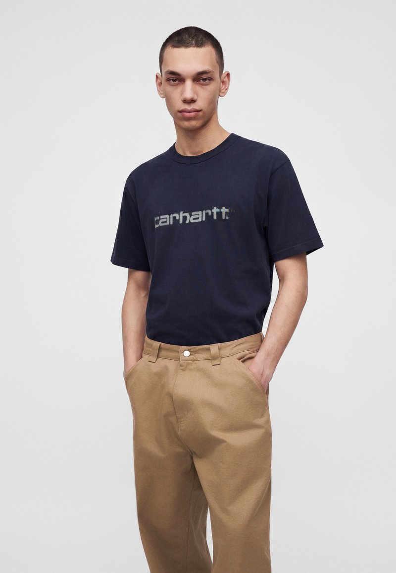 Jeune homme aux cheveux courts portant un T-shirt Carhartt bleu marine et un pantalon beige, les mains dans les poches, debout devant un fond uni.