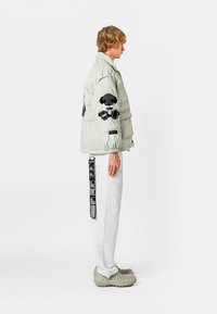 Chaqueta acolchada gris claro con parches bordados en negro, dos bolsillos frontales y puños ajustables. Usada con pantalones blancos y zapatos grises texturizados.