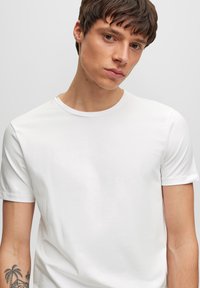 Camiseta de corte clásico, de color blanco, hecha de algodón suave. Mangas cortas con una textura lisa y un diseño minimalista. Sin logotipos ni patrones visibles.
