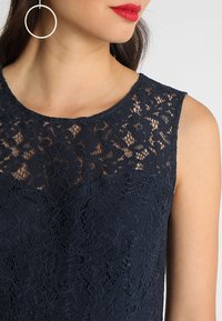 Granatowa koronkowa bluzka bez rękawów z kwiatowymi wzorami i okrągłym dekoltem. Gładka faktura materiału z przezroczystymi koronkowymi akcentami.