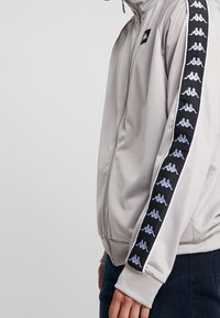 Lys grå zip-up jakke med høj krave, glat tekstur og sorte sidelister med gentaget hvidt logomønster.