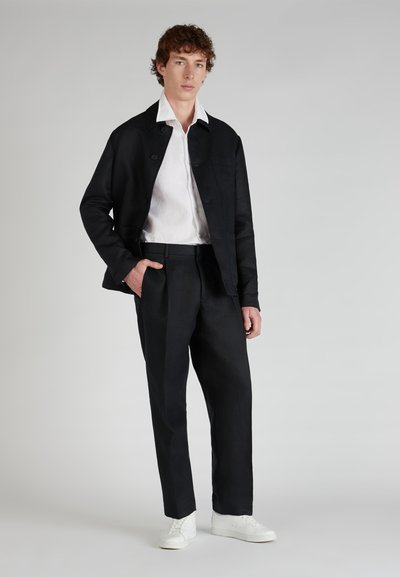Black linen jacket with a button closure and pocket, paired with pleated dark trousers and white sneakers, set against a neutral background.