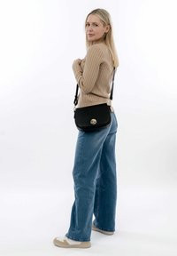 Borsa a tracolla nera con finitura texturizzata, accessori dorati e chiusura circolare. Indossata con jeans a zampa d'elefante blu chiaro e un maglione beige a coste.