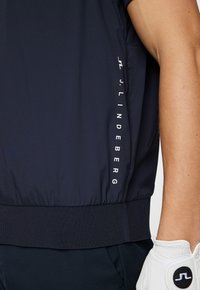 Mörkblå jacka med ribbat nederkant och sidodetaljer med "J. LINDEBERG" i vit text. Slät yta med atletisk passform.