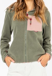 Mujer vestida con una chaqueta de forro polar verde con un bolsillo con cremallera rosa en el pecho y las manos en los bolsillos de pantalones blancos, mostrada de los hombros a las caderas.