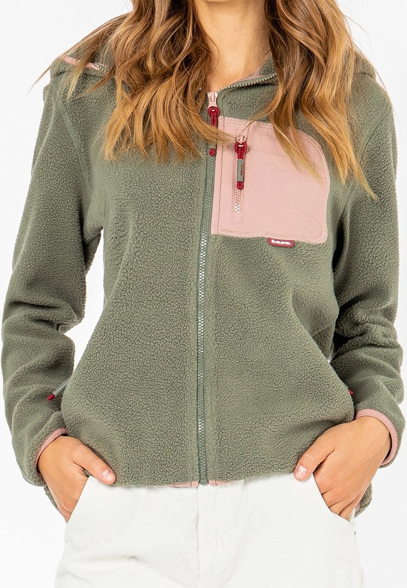 Mujer vestida con una chaqueta de forro polar verde con un bolsillo con cremallera rosa en el pecho y las manos en los bolsillos de pantalones blancos, mostrada de los hombros a las caderas.