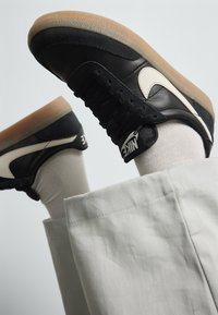 Baskets en cuir noir avec un revêtement en daim beige, semelle en caoutchouc et logo blanc ; associées à un pantalon gris clair et des chaussettes crème.