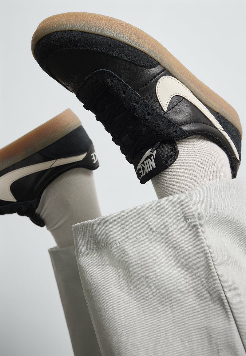 Baskets en cuir noir avec un revêtement en daim beige, semelle en caoutchouc et logo blanc ; associées à un pantalon gris clair et des chaussettes crème.