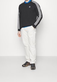 Man som står iklädd en svart Adidas-luvtröja med vita ränder, en blå polo, vita byxor och svarta sneakers med orange detaljer.