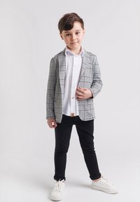 Chłopiec stoi, ubrany w białą koszulę, czarne spodnie, białe sneakersy i szary w kratkę żakiet, na tle gładkiego białego tła.