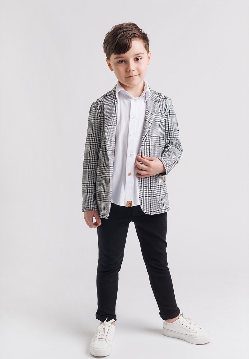 Chłopiec stoi, ubrany w białą koszulę, czarne spodnie, białe sneakersy i szary w kratkę żakiet, na tle gładkiego białego tła.