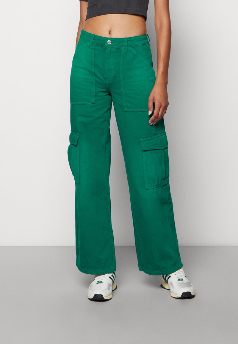 Personne portant un pantalon cargo vert à jambes larges et des baskets blanches, debout devant un fond clair uni.