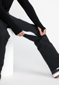 Black pants with zippers at the lower legs, showcasing a white polka-dotted inner lining. Smooth fabric, slightly loose fit, sitting pose.