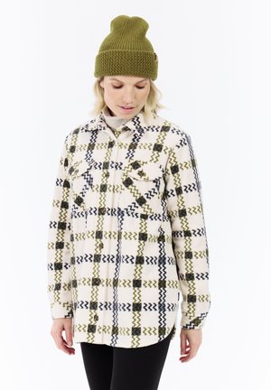 Woman wearing green knit beanie and cream shirt jacket with green and black zigzag plaid pattern, looking down, standing against white background.