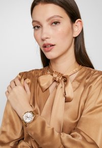 Montre en or avec un cadran blanc rond, des marqueurs argentés et un bracelet texturé en or. La personne qui la porte porte un chemisier en satin marron avec un détail de nœud.