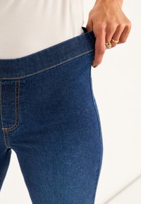 T tumma sininen farkkuhousut, joissa on leveä vyötärö, kultaiset ompeleet ja yhdistelmä sileitä ja teksturoituja pintoja.