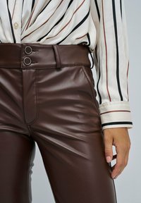 Close-up van een persoon die hoge bruine leren broek draagt met dubbele knoopsluiting en een crème blouse met zwarte en bruine verticale strepen.