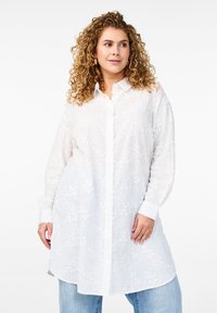 Weißes Hemd mit floraler Stickerei, langen Ärmeln und lockerer Passform, kombiniert mit hellblauen Jeans.