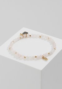 Bracelet en perles de quartz rose avec des accents dorés, comprenant un pendentif en forme de cœur et une petite étiquette. Texture lisse et couleur rose pastel.