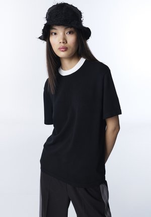 Jeune femme portant un chapeau bob noir texturé, une chemise noire à manches courtes avec col blanc, et un pantalon noir à rayures grises verticales.