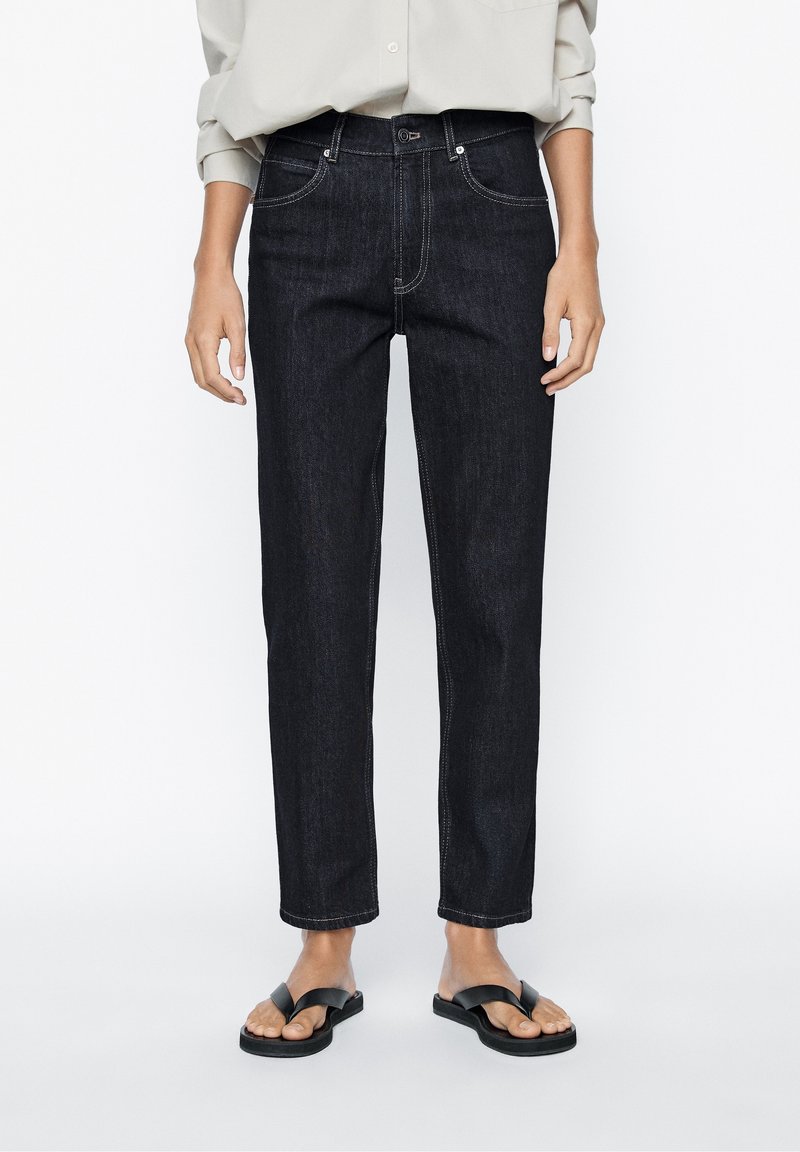 Personne portant un jean droit foncé, une chemise claire à manches longues et des sandales noires à bouts ouverts, debout devant un fond blanc.