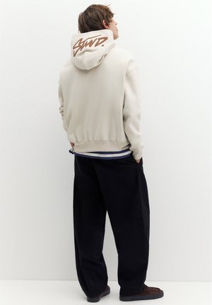 Jeune personne portant un sweat à capuche beige avec un texte marron sur la capuche, un pantalon large sombre et des chaussures foncées, debout les mains dans les poches face à un mur blanc.