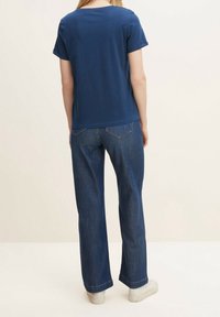 Mörkblå t-shirt med kort ärm och rund halsringning, tillsammans med vida denimbyxor och vita sneakers, sett bakifrån.