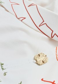 Biely textil s červenými a zelenými abstraktnými líniovými vzormi, obsahujúci malú ozdobnú zlatú sponu s prepracovanými zdobenými motívmi.