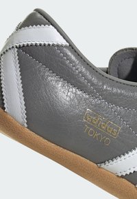 Γκρι αθλητικό παπούτσι Adidas Tokyo με λευκές ζιγκ ζαγκ ρίγες και σόλα από καουτσούκ καφέ, που παρουσιάζει υφή δέρματος και ραμμένες λεπτομέρειες.