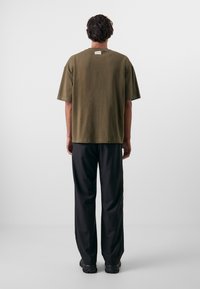 Oliven grøn bomulds T-shirt med korte ærmer og lige kant, båret med sorte bukser og sort fodtøj, set fra ryggen.