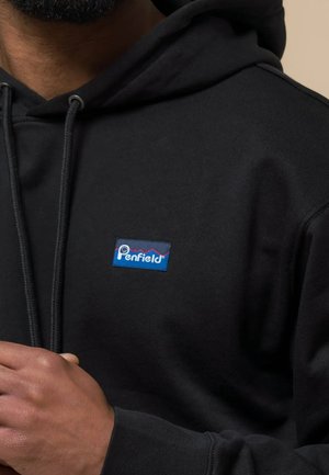 Persona che indossa una felpa con cappuccio nera Penfield con patch logo frontale e coulisse, mano parzialmente visibile vicino al petto.