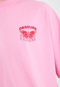 Rosa Baumwoll-T-Shirt mit rotem Text "OBSCURE FUTURE" und einem Schmetterlingsgrafik. Weicher Stoff, Rundhalsausschnitt, kurze Ärmel.