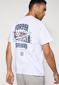 T-shirt branca de algodão exibindo um gráfico nas costas com a frase "FOREVER FORWARD", tema de futebol, realces em azul e vermelho, e estrelas.