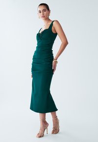 Teal fitted dress with a scooped neckline, ruched detailing, and a midi length, paired with gold strappy heels. Smooth, textured fabric.