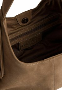 Interno della borsa in suede marrone, con una tasca con zip e una patch con il logo. La fodera è di un marrone più scuro con cuciture visibili. Texture liscia.