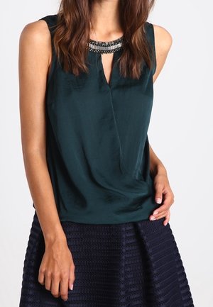 Mujer con blusa sin mangas verde oscuro con escote con cuentas y falda azul marino texturizada, con la mano apoyada en la cadera.