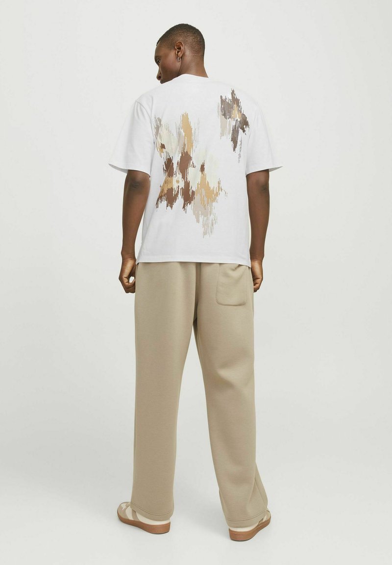 Witte t-trui met korte mouwen met een abstract bruin en beige patroon op de achterkant; gecombineerd met beige broek en lichtbruine schoenen.